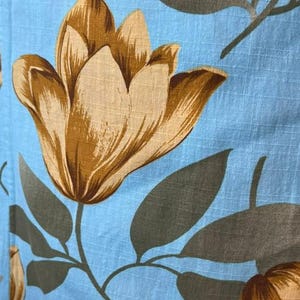 May include: A light blue fabric featuring a repeating pattern of large, tan-colored tulip-like flowers and dark gray leaves. The textile has a visible grid-like texture throughout, creating a subtle checked appearance across the floral design.