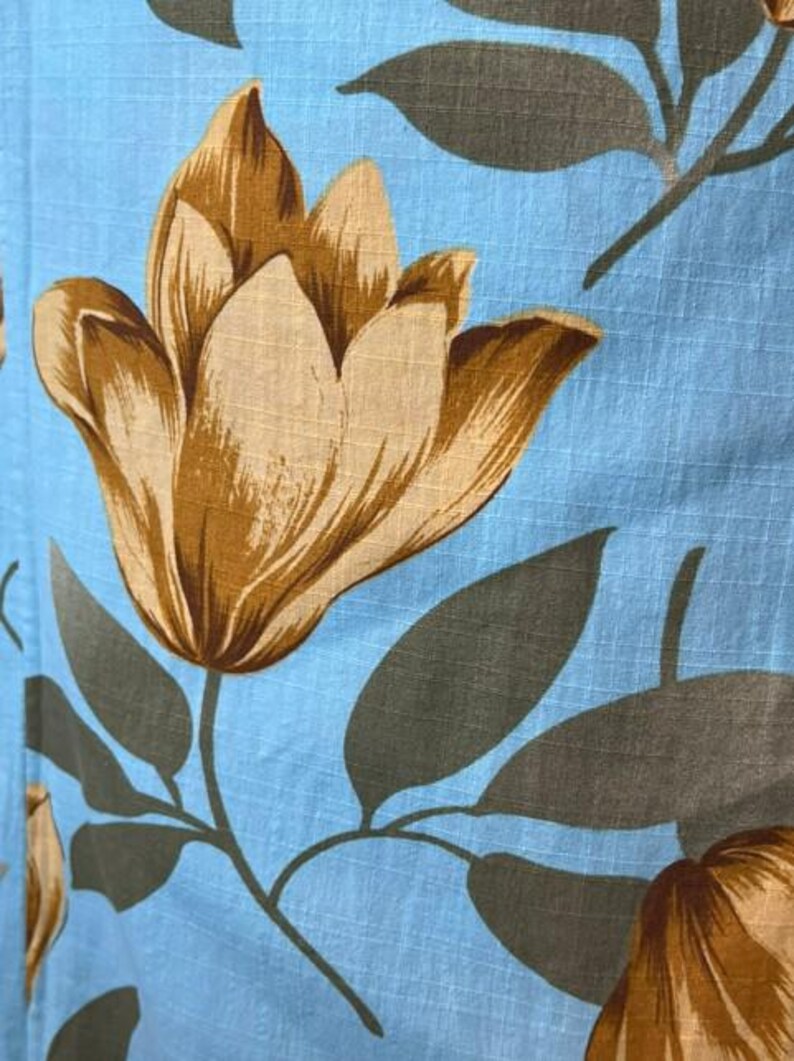 May include: A light blue fabric featuring a repeating pattern of large, tan-colored tulip-like flowers and dark gray leaves. The textile has a visible grid-like texture throughout, creating a subtle checked appearance across the floral design.