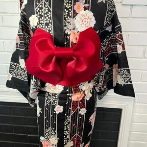 May include: A black kimono featuring a floral pattern with white and pink blossoms, accented by vertical geometric lines. A large, vibrant red satin bow is secured at the waist, contrasting against the dark fabric of the garment.