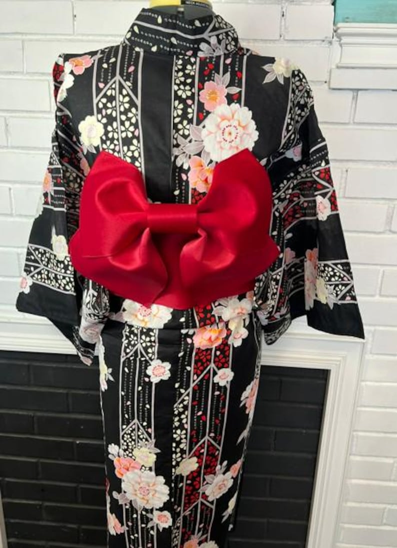 May include: A black kimono featuring a floral pattern with white and pink blossoms, accented by vertical geometric lines. A large, vibrant red satin bow is secured at the waist, contrasting against the dark fabric of the garment.