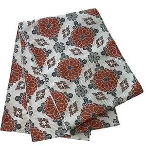 May include: A folded piece of fabric featuring a white background with a repeating pattern of rust-coloured chrysanthemum flowers, small grey floral accents, and geometric diamond-shaped motifs arranged in a structured, symmetrical layout.