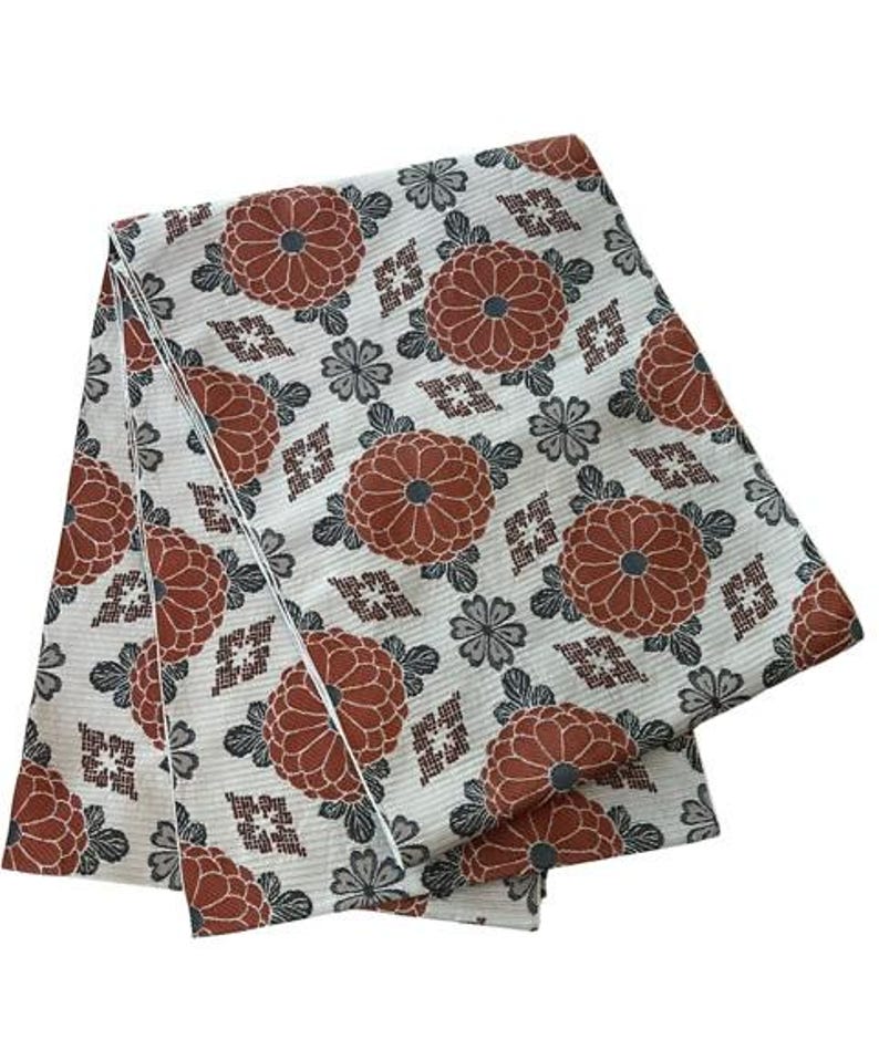 May include: A folded piece of fabric featuring a white background with a repeating pattern of rust-coloured chrysanthemum flowers, small grey floral accents, and geometric diamond-shaped motifs arranged in a structured, symmetrical layout.