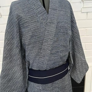 Może przedstawiać: Kimono w ciemnoniebiesko-biały wz&oacute;r z drobną, powtarzającą się geometryczną teksturą na całej tkaninie. Ubi&oacute;r posiada szerokie rękawy, tradycyjny dekolt typu kopertowego i jest przewiązany w talii gładkim, granatowym pasem typu obi.
