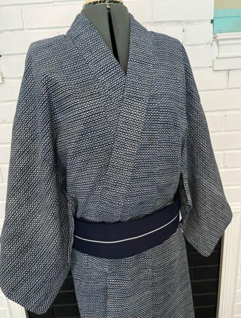 Może przedstawiać: Kimono w ciemnoniebiesko-biały wz&oacute;r z drobną, powtarzającą się geometryczną teksturą na całej tkaninie. Ubi&oacute;r posiada szerokie rękawy, tradycyjny dekolt typu kopertowego i jest przewiązany w talii gładkim, granatowym pasem typu obi.