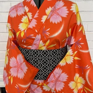 Może przedstawiać: Pomarańczowe kimono z powtarzającym się wzorem r&oacute;żowych i ż&oacute;łtych kwiat&oacute;w wiśni oraz małych fioletowych ważek. Ubi&oacute;r posiada szeroki, czarny pas obi z białym geometrycznym wzorem kratki, długie rękawy oraz zapięcie typu kopertowego.