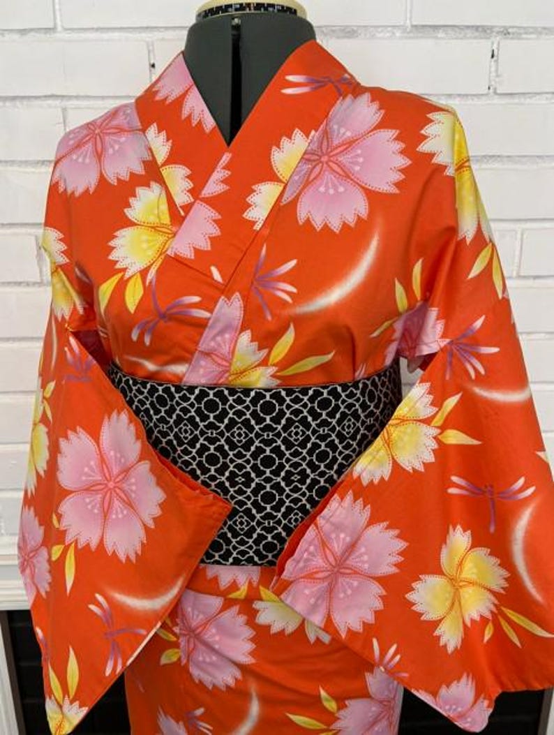 Może przedstawiać: Pomarańczowe kimono z powtarzającym się wzorem r&oacute;żowych i ż&oacute;łtych kwiat&oacute;w wiśni oraz małych fioletowych ważek. Ubi&oacute;r posiada szeroki, czarny pas obi z białym geometrycznym wzorem kratki, długie rękawy oraz zapięcie typu kopertowego.