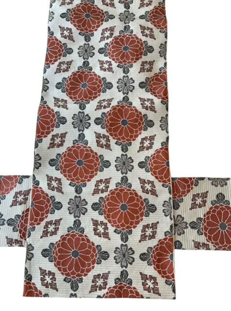 May include: A textile fabric featuring a repeating pattern of large, rust-orange circular floral motifs interspersed with smaller grey flower shapes and geometric diamond-like accents on a cream-coloured background with fine horizontal lines.