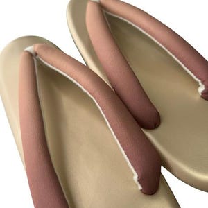 May include: A pair of traditional Japanese geta sandals. The sandals have a light tan base with a curved design and a dark pink thong. The thong is made of a soft material and is attached to the base with a white trim.