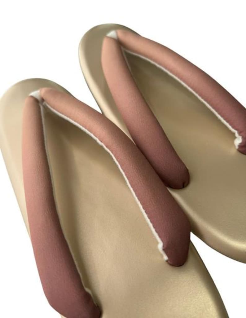 May include: A pair of traditional Japanese geta sandals. The sandals have a light tan base with a curved design and a dark pink thong. The thong is made of a soft material and is attached to the base with a white trim.