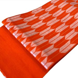 May include: A vibrant orange fabric featuring a repeating white arrow-feather pattern. The textile is folded to display both a solid orange section and a section with the geometric arrow motif, showcasing a consistent weave and bright color.