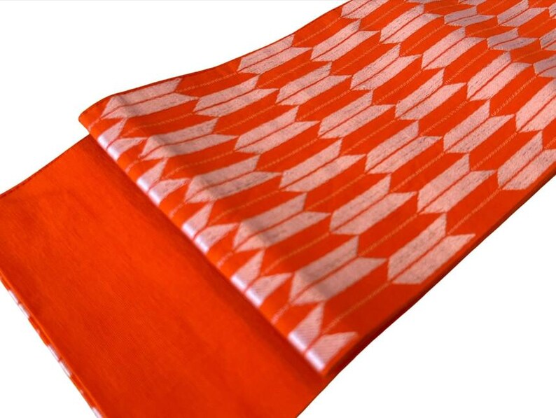 May include: A vibrant orange fabric featuring a repeating white arrow-feather pattern. The textile is folded to display both a solid orange section and a section with the geometric arrow motif, showcasing a consistent weave and bright color.