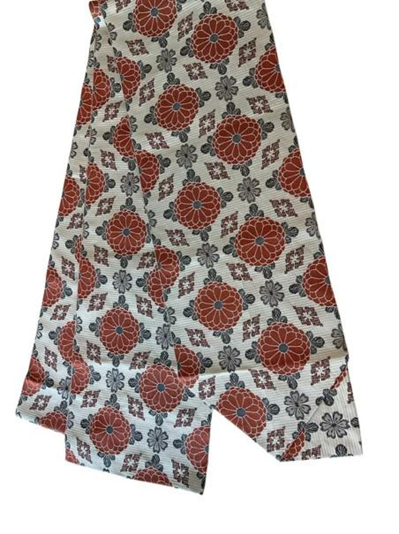 May include: A long, rectangular textile strip featuring a repeating pattern of large, rust-orange chrysanthemum-style floral motifs interspersed with smaller, dark grey floral shapes and geometric diamond-like accents on a cream-coloured background.