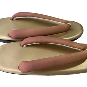 May include: A pair of traditional Japanese geta sandals. The sandals have a light tan base with pink straps and white padding. The soles are black. The geta are designed for outdoor wear and are elevated.