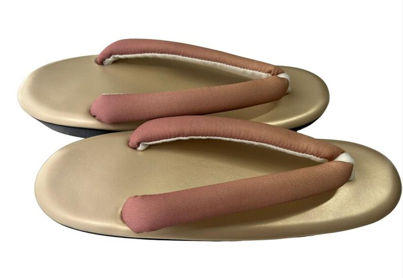 May include: A pair of traditional Japanese geta sandals. The sandals have a light tan base with pink straps and white padding. The soles are black. The geta are designed for outdoor wear and are elevated.