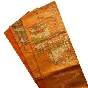 Op de afbeelding: Een oranje textiel met een geometrisch patroon en decoratieve doosontwerpen in tinten groen, goud en rood. De stof is gevouwen en onthult een effen oranje rand. Het algehele ontwerp suggereert een traditionele of vintage stijl.