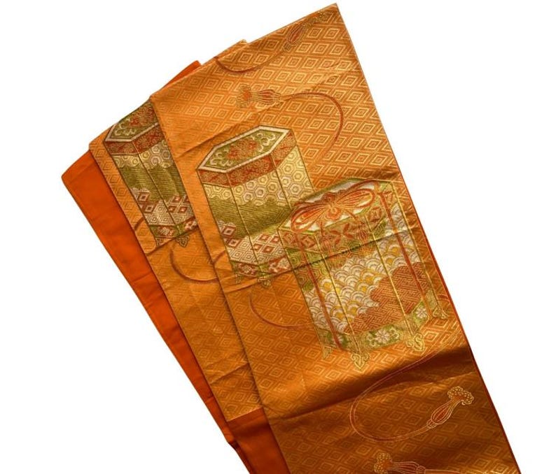 Op de afbeelding: Een oranje textiel met een geometrisch patroon en decoratieve doosontwerpen in tinten groen, goud en rood. De stof is gevouwen en onthult een effen oranje rand. Het algehele ontwerp suggereert een traditionele of vintage stijl.