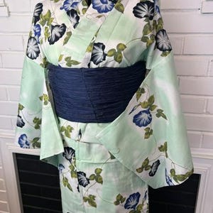 Op de afbeelding: Een licht mintgroene kimono met een herhalend patroon van blauwe en witte ochtendgloriebloemen met groene bladeren. Het kledingstuk is in de taille ingesnoerd met een brede, getextureerde, donkerblauwe obi-ceintuur voor een gestructureerd silhouet.