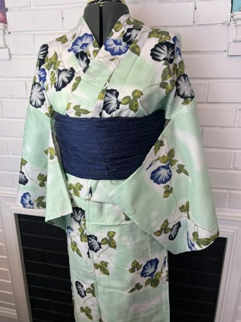 Op de afbeelding: Een licht mintgroene kimono met een herhalend patroon van blauwe en witte ochtendgloriebloemen met groene bladeren. Het kledingstuk is in de taille ingesnoerd met een brede, getextureerde, donkerblauwe obi-ceintuur voor een gestructureerd silhouet.