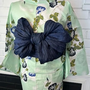 Op de afbeelding: Een licht muntgroene yukata met een herhalend patroon van blauwe en witte ochtendgloriebloemen met groene bladeren. Het kledingstuk is in de taille gestrikt met een grote, getextureerde, donker marineblauwe obi-sjerp in een strikvorm.