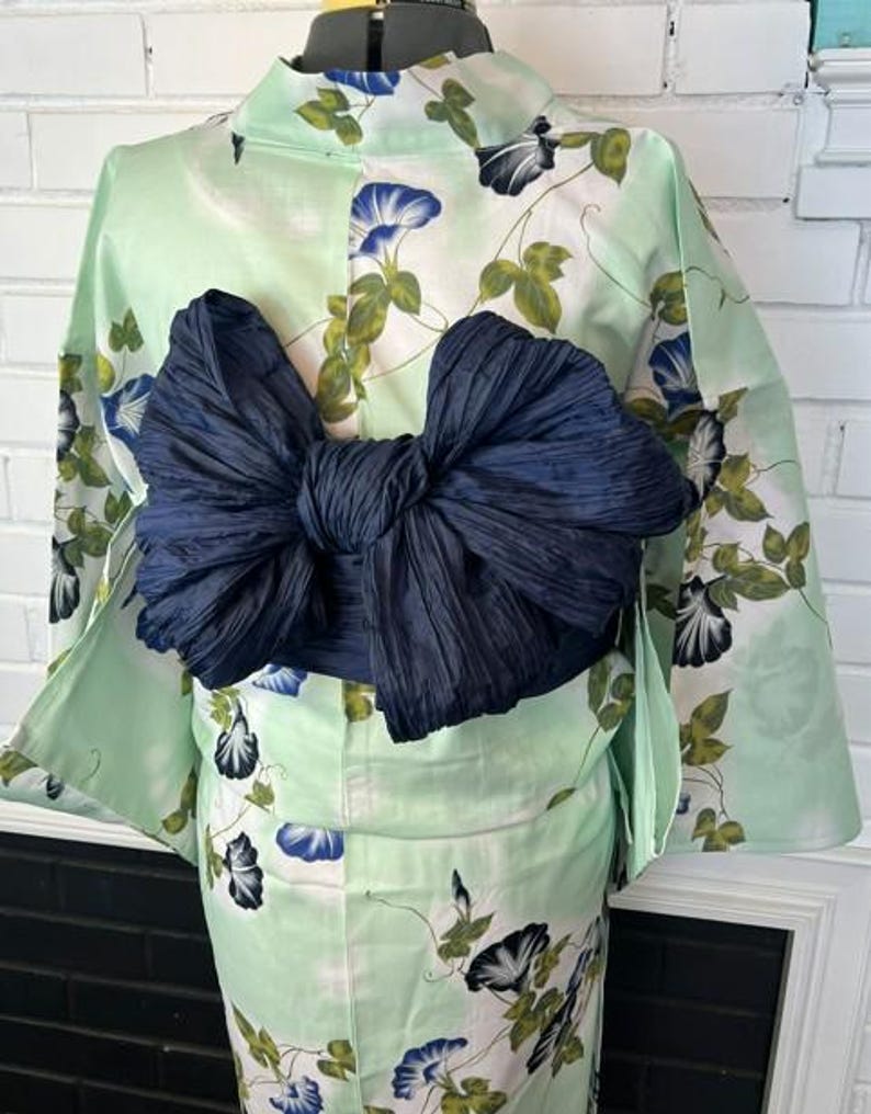 Op de afbeelding: Een licht muntgroene yukata met een herhalend patroon van blauwe en witte ochtendgloriebloemen met groene bladeren. Het kledingstuk is in de taille gestrikt met een grote, getextureerde, donker marineblauwe obi-sjerp in een strikvorm.