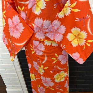 Może przedstawiać: Jaskrawopomarańczowe kimono z powtarzającym się wzorem dużych r&oacute;żowych i ż&oacute;łtych kwiat&oacute;w wiśni. Ubi&oacute;r posiada szerokie, luźne rękawy i jest ozdobiony małymi, delikatnymi ilustracjami fioletowych ważek rozmieszczonymi na całej tkaninie.