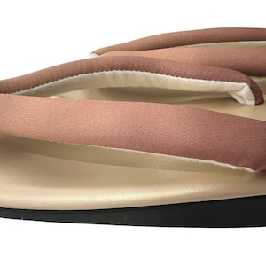 May include: Close-up of a pair of traditional Japanese geta sandals. The sandals feature a light tan base with a black sole and three straps in a muted rose color. The straps are padded with a white interior.