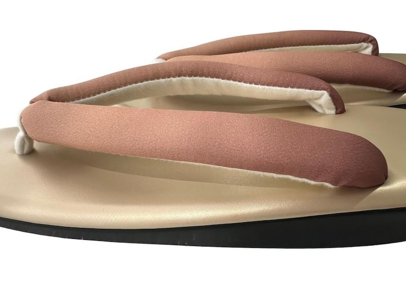 May include: Close-up of a pair of traditional Japanese geta sandals. The sandals feature a light tan base with a black sole and three straps in a muted rose color. The straps are padded with a white interior.