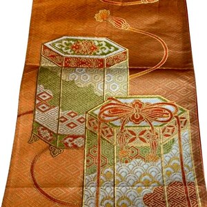 Op de afbeelding: Een oranje textiel met ingewikkelde patronen en twee decoratieve, geometrische vormen. De vormen zijn goudkleurig omlijnd en hebben rode, groene en witte details. Fijne gouden lijnen en bloemaccenten verfraaien het ontwerp.