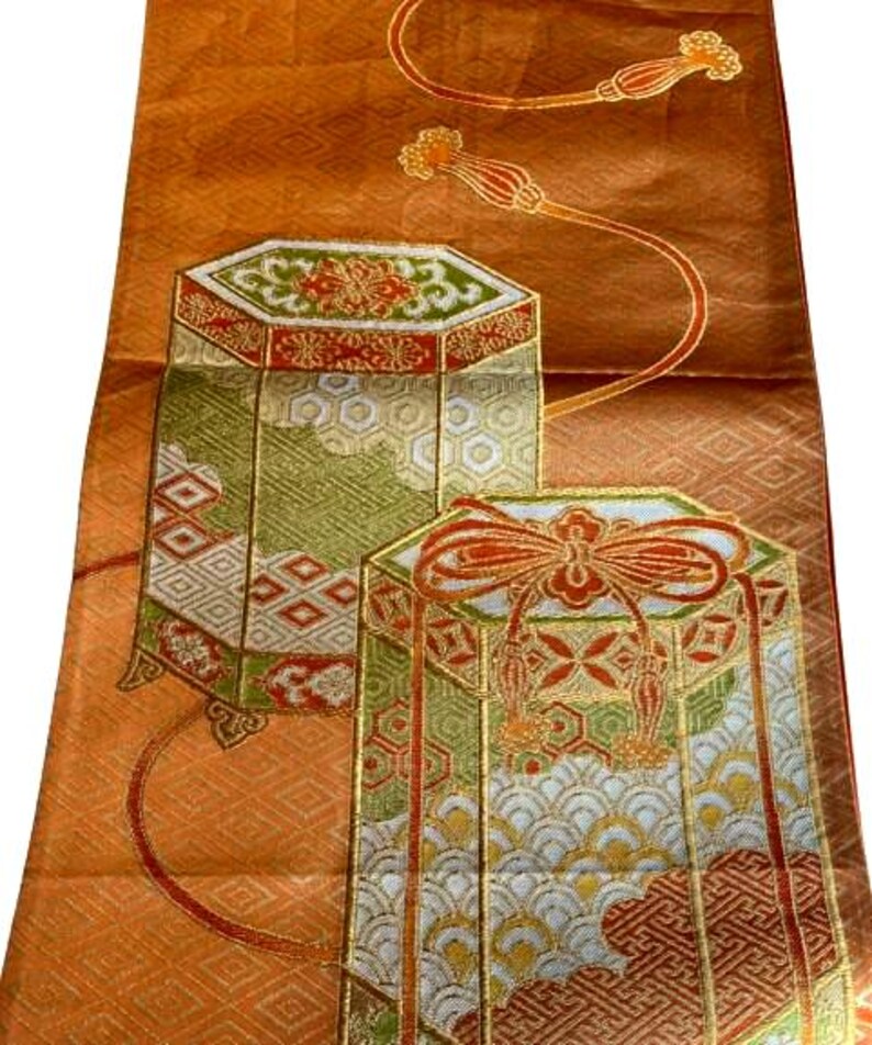 Op de afbeelding: Een oranje textiel met ingewikkelde patronen en twee decoratieve, geometrische vormen. De vormen zijn goudkleurig omlijnd en hebben rode, groene en witte details. Fijne gouden lijnen en bloemaccenten verfraaien het ontwerp.