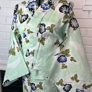 Op de afbeelding: Een licht mintgroene kimono met een herhalend patroon van blauwe en witte ochtendgloriebloemen met groene bladeren en dunne bruine ranken. Het kledingstuk heeft wijde mouwen, een overslagsluiting en een gestructureerde kraag op een paspop.