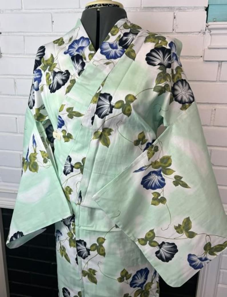Op de afbeelding: Een licht mintgroene kimono met een herhalend patroon van blauwe en witte ochtendgloriebloemen met groene bladeren en dunne bruine ranken. Het kledingstuk heeft wijde mouwen, een overslagsluiting en een gestructureerde kraag op een paspop.