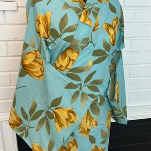 May include: A light blue kimono featuring a repeating pattern of large golden yellow flowers and muted olive green leaves. The garment has wide sleeves, a wrap-style front with a V-neckline, and a structured collar, displayed on a mannequin torso.