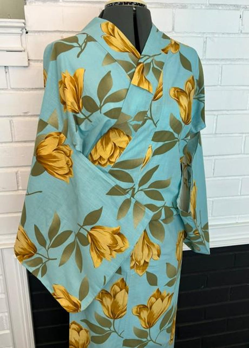 May include: A light blue kimono featuring a repeating pattern of large golden yellow flowers and muted olive green leaves. The garment has wide sleeves, a wrap-style front with a V-neckline, and a structured collar, displayed on a mannequin torso.