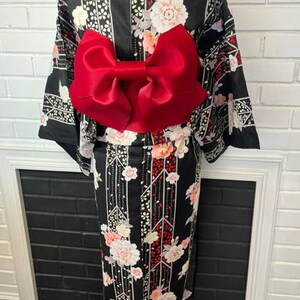 May include: A black kimono featuring a floral pattern with white and pink blossoms, geometric lines, and a large, solid red bow tied at the waist. The garment has long, wide sleeves and a high collar, displayed on a mannequin form.