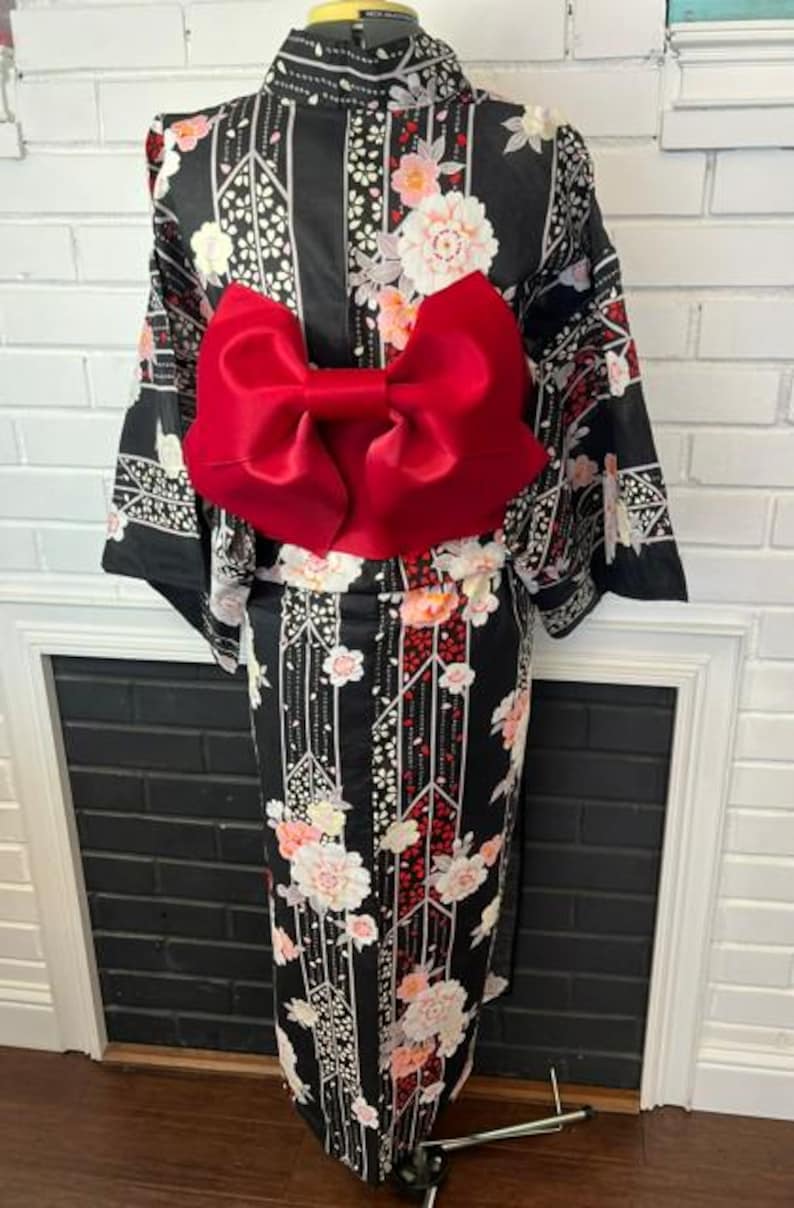 May include: A black kimono featuring a floral pattern with white and pink blossoms, geometric lines, and a large, solid red bow tied at the waist. The garment has long, wide sleeves and a high collar, displayed on a mannequin form.