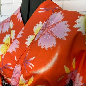 Może przedstawiać: Pomarańczowe kimono z powtarzającym się wzorem dużych r&oacute;żowych i ż&oacute;łtych kwiat&oacute;w wiśni oraz małych fioletowych ważek. Odzież posiada dekolt w serek z szerokim kołnierzem i jest zaprezentowana na manekinie, aby ukazać nadruk na tkaninie.