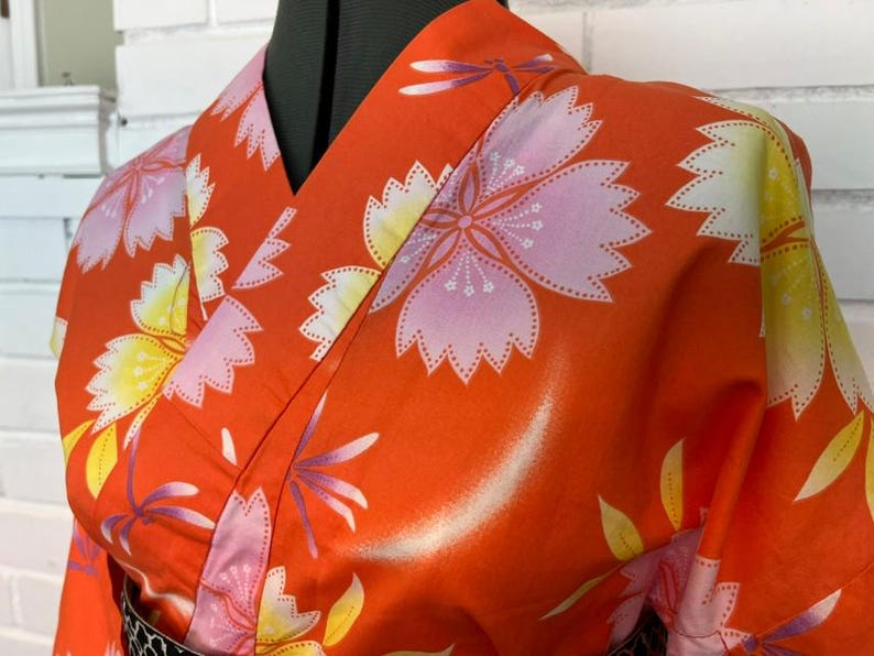 Może przedstawiać: Pomarańczowe kimono z powtarzającym się wzorem dużych r&oacute;żowych i ż&oacute;łtych kwiat&oacute;w wiśni oraz małych fioletowych ważek. Odzież posiada dekolt w serek z szerokim kołnierzem i jest zaprezentowana na manekinie, aby ukazać nadruk na tkaninie.