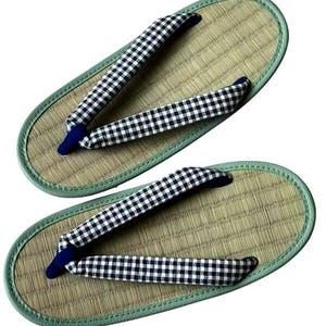 May include: Pair of traditional Japanese geta sandals. The sandals feature a woven straw base with a light green border. The straps are black and white checkered fabric with a navy blue toe post. The geta are designed for outdoor wear.
