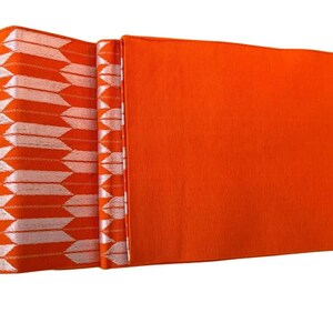 May include: A vibrant orange obi sash featuring a repeating white arrow feather pattern on one end. The fabric displays a smooth, solid orange section alongside a textured segment with white arrow motifs arranged in vertical columns.
