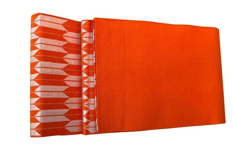 May include: A vibrant orange obi sash featuring a repeating white arrow feather pattern on one end. The fabric displays a smooth, solid orange section alongside a textured segment with white arrow motifs arranged in vertical columns.