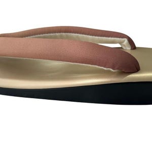 May include: A traditional Japanese geta sandal. The sandal features a gold-colored platform with a black base. The thong strap is a muted rose color with a white and beige lining. The sandal is designed for comfort and style.