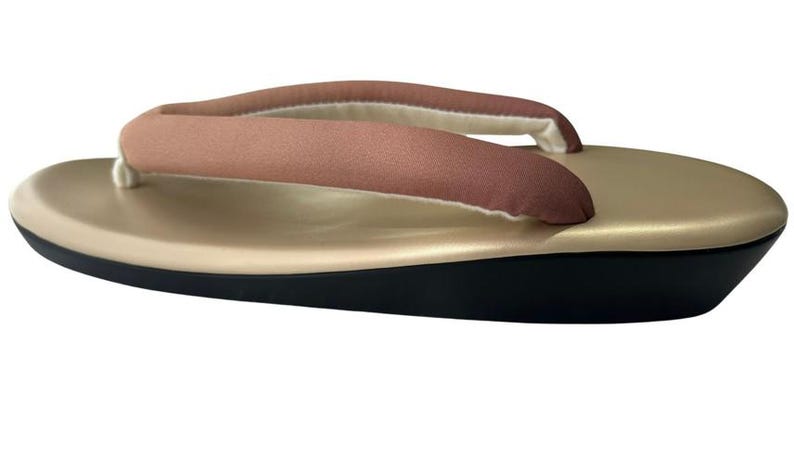 May include: A traditional Japanese geta sandal. The sandal features a gold-colored platform with a black base. The thong strap is a muted rose color with a white and beige lining. The sandal is designed for comfort and style.