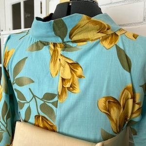 May include: A light blue blouse featuring a high neckline and a repeating floral pattern in shades of golden yellow and muted olive green. The garment is displayed on a dress form, showcasing long sleeves and a structured collar design.