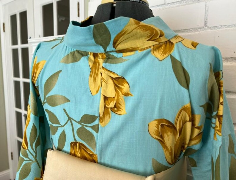 May include: A light blue blouse featuring a high neckline and a repeating floral pattern in shades of golden yellow and muted olive green. The garment is displayed on a dress form, showcasing long sleeves and a structured collar design.