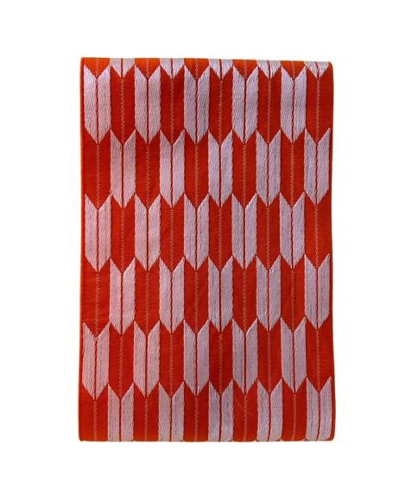 May include: A rectangular textile piece featuring a vibrant orange background with a repeating pattern of light pink arrow-like shapes arranged in vertical columns. The fabric displays a consistent woven texture throughout the entire surface area.
