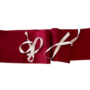 May include: A long, rectangular, deep red fabric wrap featuring two sets of thin, off-white ribbon ties attached to the front. The fabric has a smooth, matte texture and is laid out horizontally, showing the ties loosely draped over the surface.