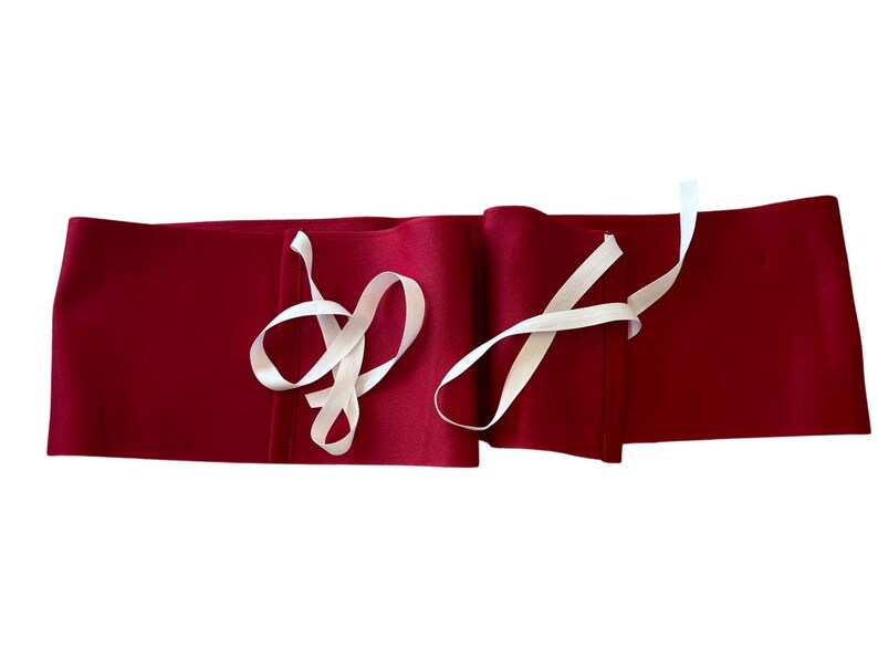 May include: A long, rectangular, deep red fabric wrap featuring two sets of thin, off-white ribbon ties attached to the front. The fabric has a smooth, matte texture and is laid out horizontally, showing the ties loosely draped over the surface.