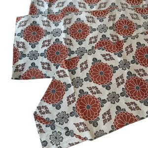 May include: A white fabric runner featuring a repeating pattern of large terracotta-coloured chrysanthemum flowers, smaller grey floral motifs, and geometric diamond shapes. The textile has a fine, vertical ribbed texture throughout the surface.