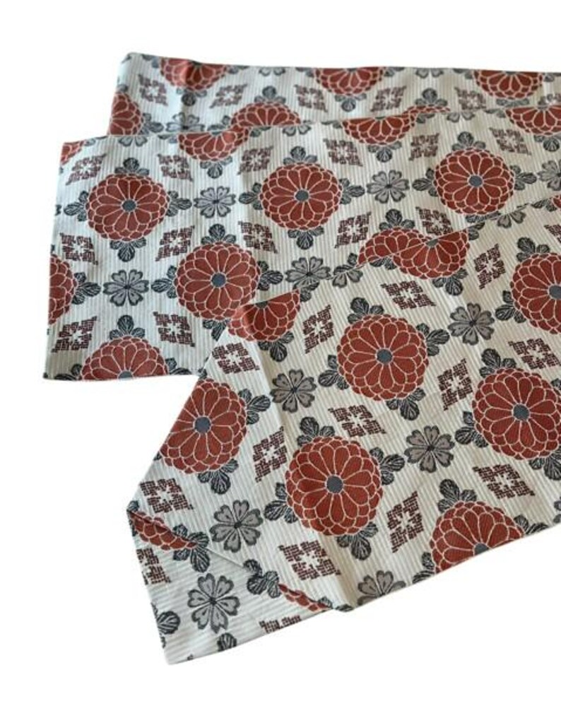 May include: A white fabric runner featuring a repeating pattern of large terracotta-coloured chrysanthemum flowers, smaller grey floral motifs, and geometric diamond shapes. The textile has a fine, vertical ribbed texture throughout the surface.