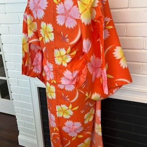 Może przedstawiać: Jasnopomarańczowe yukata z powtarzającym się kwiatowym wzorem r&oacute;żowych i ż&oacute;łtych kwiat&oacute;w wiśni. Ubi&oacute;r posiada szerokie rękawy, wysoki kołnierz oraz prosty kr&oacute;j, prezentując wyrazisty botaniczny nadruk na całej powierzchni tkaniny.